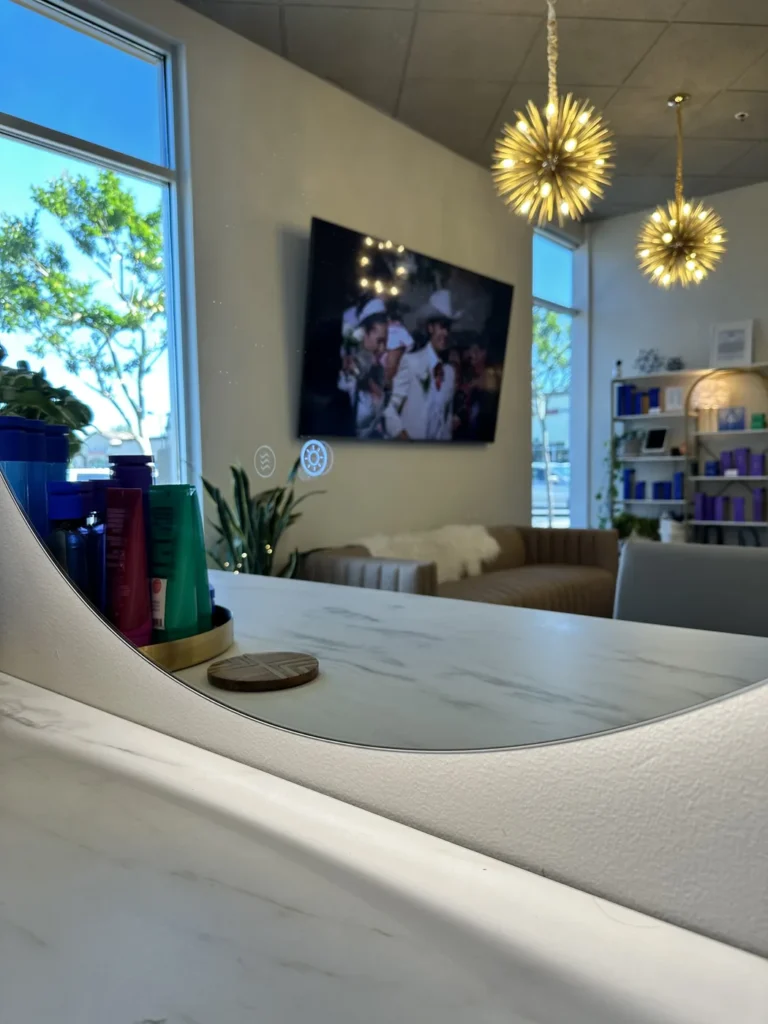 Lounge and reception at Vamp Salon Extension Studio, a hair salon in Brentwood, CA, with gold sputnik chandeliers, a marble counter, and front windows