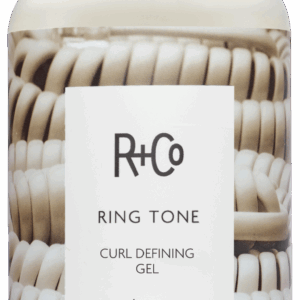 Bottle of R+Co Ring Tone Curl Defining Gel with white label, beige cap, and stacked beige coils in the background; reads Ring Tone Curl Defining Gel 177 ml / 6 fl oz.